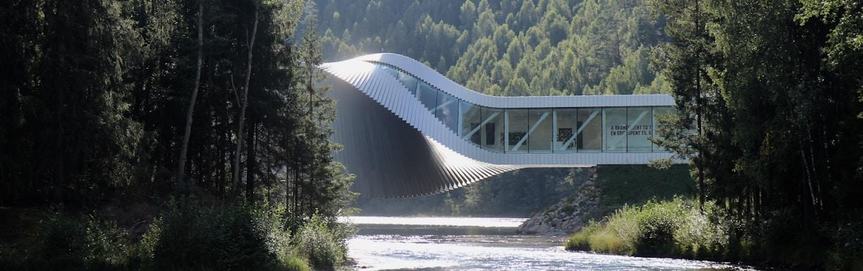 An innovative architectural structure stretches across a river, blending harmoniously with the lush, forested landscape. The building has a dynamic, twisting form, which combines modern design with the natural environment. Sunlight reflects off the surface of the river, creating a serene and tranquil atmosphere amidst the tall trees that flank both sides.