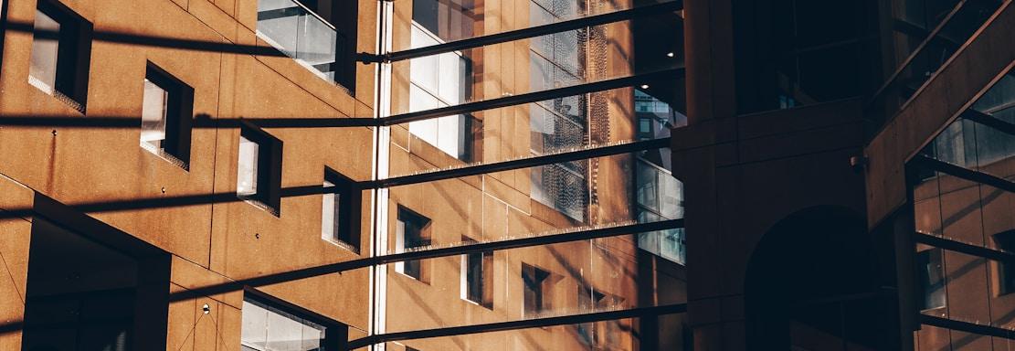 A modern architectural structure with large glass windows and metal frames allowing sunlight to cast intricate shadows on the walls and floor. The interior showcases a sophisticated design with a warm color palette created by the sunlight.