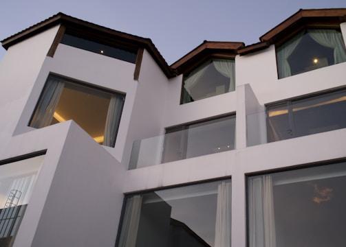 Contemporary architecture featuring a white multi-level building with large windows and dark slanted roofs. The structure is modern and minimalistic, exuding a sense of sophistication. Glass balconies and drapes can also be seen through the expansive windows.