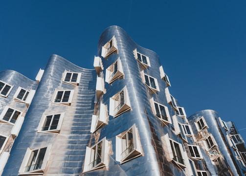 An architectural structure with a modern, innovative design featuring a wavy facade made of reflective metallic panels. Numerous windows with open shutters are distributed unevenly across the building's surface. The structure is set against a clear blue sky.